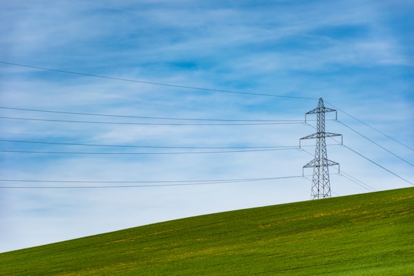 A long distance power transmission line in front of a blue sky with light white clouds, a green field in front.