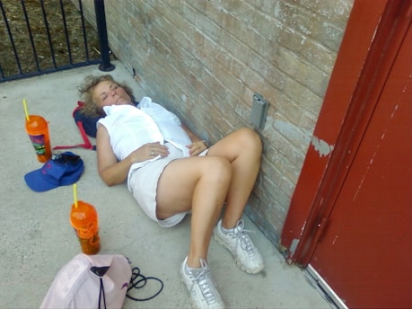 A person sleeping or resting against a brick wall on a concrete floor, wearing white clothing and sneakers. Two large orange cups with straws are nearby, along with a blue cap and a pink bag.
