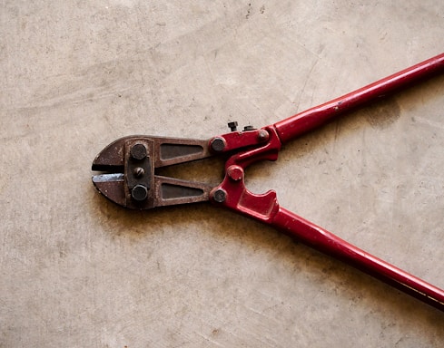 A pair of red bolt cutters rests on a concrete surface. The metal jaws and pivot mechanism are visible, showcasing the tool's sturdy construction.