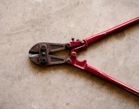 A pair of red bolt cutters rests on a concrete surface. The metal jaws and pivot mechanism are visible, showcasing the tool's sturdy construction.