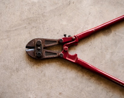 A pair of red bolt cutters rests on a concrete surface. The metal jaws and pivot mechanism are visible, showcasing the tool's sturdy construction.