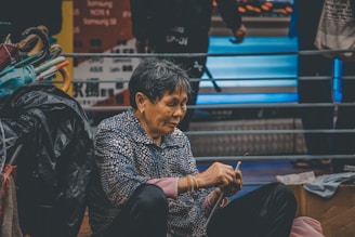 An older person with short hair is sitting on the ground, holding fabric or thread, and appears to be sewing or mending something. To the left are colorful umbrellas and other belongings. The background features a railing and blurred signs or advertisements with text.