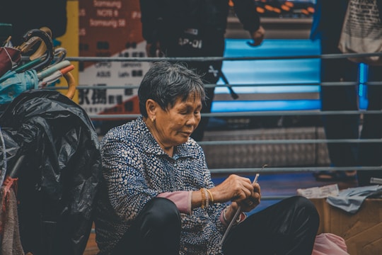 An older person with short hair is sitting on the ground, holding fabric or thread, and appears to be sewing or mending something. To the left are colorful umbrellas and other belongings. The background features a railing and blurred signs or advertisements with text.