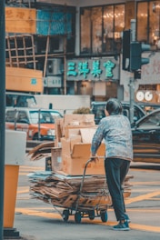 A person is pushing a cart loaded with cardboard boxes and flattened cardboard through a busy street intersection. The background features a vibrant urban setting with multiple cars and storefronts displaying bright signs in Chinese characters.