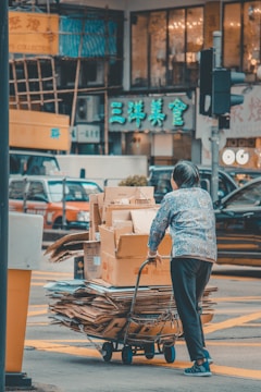 A person is pushing a cart loaded with cardboard boxes and flattened cardboard through a busy street intersection. The background features a vibrant urban setting with multiple cars and storefronts displaying bright signs in Chinese characters.