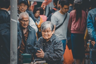 A bustling street scene with a crowd of people, prominently featuring an elderly woman with short gray hair wearing glasses and a patterned jacket. She stands amidst other individuals of various ages, some looking in different directions, creating a sense of movement and busyness.