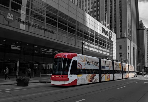 A modern red and white streetcar travels down a city street in front of the Bank of Montreal building. The streetcar is adorned with colorful advertisements featuring words like 'Savour' and images of food. The background is black and white, highlighting the vibrant streetcar.