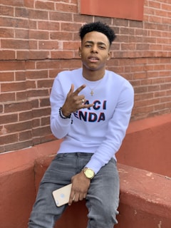 A young person is sitting against a brick wall, wearing a white sweatshirt with text, jeans, and a watch. They are holding a smartphone and making a hand gesture, possibly a peace sign or similar. Their expression appears calm and relaxed.