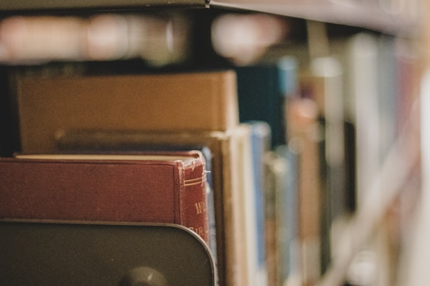 Books on a shelf with a focus on the spine of a red hardcover book, surrounded by other books with varying colors, in a library or study setting.