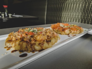 A rectangular white plate with two gourmet dishes featuring savory toppings placed on a tabletop. The dishes appear to have a crispy base with toppings including shredded vegetables, fish slices, and garnishes, drizzled with a dark sauce. Background elements include blurred tableware and condiments, adding a casual dining ambiance.