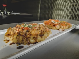 A rectangular white plate with two gourmet dishes featuring savory toppings placed on a tabletop. The dishes appear to have a crispy base with toppings including shredded vegetables, fish slices, and garnishes, drizzled with a dark sauce. Background elements include blurred tableware and condiments, adding a casual dining ambiance.