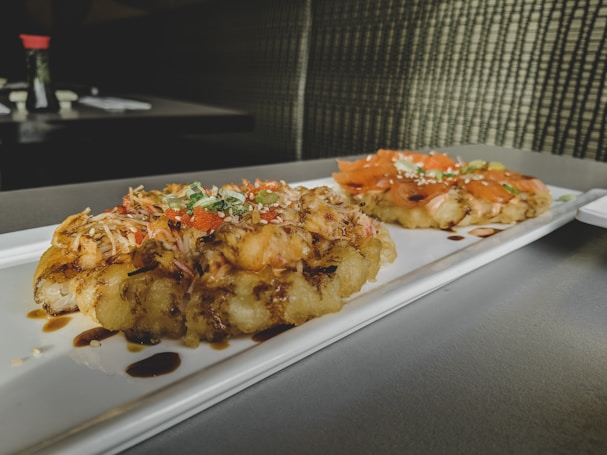 A rectangular white plate with two gourmet dishes featuring savory toppings placed on a tabletop. The dishes appear to have a crispy base with toppings including shredded vegetables, fish slices, and garnishes, drizzled with a dark sauce. Background elements include blurred tableware and condiments, adding a casual dining ambiance.