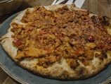 A pizza topped with generous portions of cooked chicken and red peppers, served on a stoneware plate. The base is crispy and slightly charred, with visible pieces of succulent meat and vegetables spread evenly.