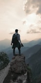 A person stands on a rocky cliff edge overlooking a vast mountain range. The person is wearing a backpack and holding a camera, suggesting exploration or photography. The sky is partly cloudy and the sun is setting or rising, casting a warm glow over the hazy mountains.