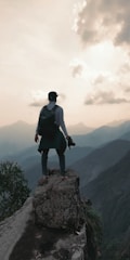 A person stands on a rocky cliff edge overlooking a vast mountain range. The person is wearing a backpack and holding a camera, suggesting exploration or photography. The sky is partly cloudy and the sun is setting or rising, casting a warm glow over the hazy mountains.