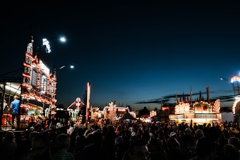 An evening fairground scene bustling with people, brightly lit attractions, and food stalls. The sky is a deep blue as night falls, and the vibrant lights illuminate the area, creating an energetic atmosphere. A large crowd moves among the various rides and stands, and the illuminated signs and decorations add a festive touch.
