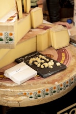 A selection of cheese is displayed, featuring several large wedges and slices of Comté cheese. Small cubes of cheese are arranged on a black slate board for sampling. The cheese wheels have distinctive labels with the Comté branding visible. A paper-wrapped block of cheese with a price written on it is also present.