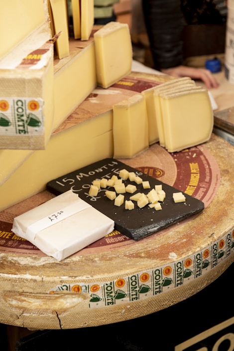 A selection of cheese is displayed, featuring several large wedges and slices of Comté cheese. Small cubes of cheese are arranged on a black slate board for sampling. The cheese wheels have distinctive labels with the Comté branding visible. A paper-wrapped block of cheese with a price written on it is also present.