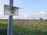 A rural landscape with a field showing rows of crops under a clear sky with scattered clouds. A metal signpost is seen on the left with 'CICLO 400' written on it. In the background, there are bushes and a few trees dotting the horizon.
