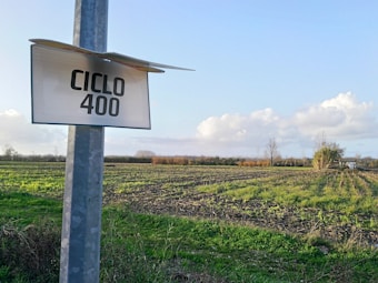 A rural landscape with a field showing rows of crops under a clear sky with scattered clouds. A metal signpost is seen on the left with 'CICLO 400' written on it. In the background, there are bushes and a few trees dotting the horizon.