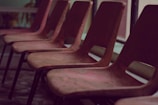 A row of empty, brown chairs lined up in an indoor setting with soft lighting. The chairs are simple in design, possibly made of plastic or wood, and are positioned in front of large windows.