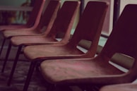 A row of empty, brown chairs lined up in an indoor setting with soft lighting. The chairs are simple in design, possibly made of plastic or wood, and are positioned in front of large windows.