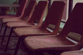 A row of empty, brown chairs lined up in an indoor setting with soft lighting. The chairs are simple in design, possibly made of plastic or wood, and are positioned in front of large windows.