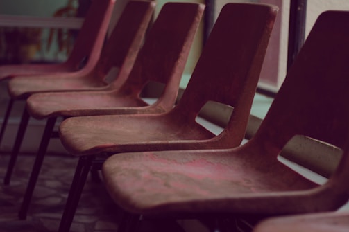 A row of empty, brown chairs lined up in an indoor setting with soft lighting. The chairs are simple in design, possibly made of plastic or wood, and are positioned in front of large windows.