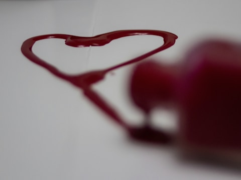 A heart shape is drawn with red liquid on a white surface, with a blurred reddish bottle in the foreground.