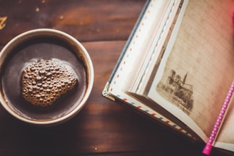 A steaming cup of black coffee sits on a wooden table next to an open notebook. The notebook features lined pages and an image of a historic building. A pink pen rests on the notebook, creating a cozy and contemplative atmosphere.