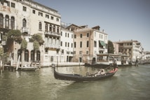 A gondola glides through a canal in Venice, Italy, with historic buildings lining the waterway. The buildings feature rustic facades and arched windows, typical of Venetian architecture. Several people are seated in the gondola, enjoying the serene ride under the clear sky.
