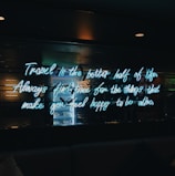 A neon sign with a motivational quote is illuminated against a dark background. The quote encourages taking time for things that bring happiness. Reflections and a slight glow from the neon text are visible.