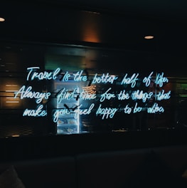 A neon sign with a motivational quote is illuminated against a dark background. The quote encourages taking time for things that bring happiness. Reflections and a slight glow from the neon text are visible.