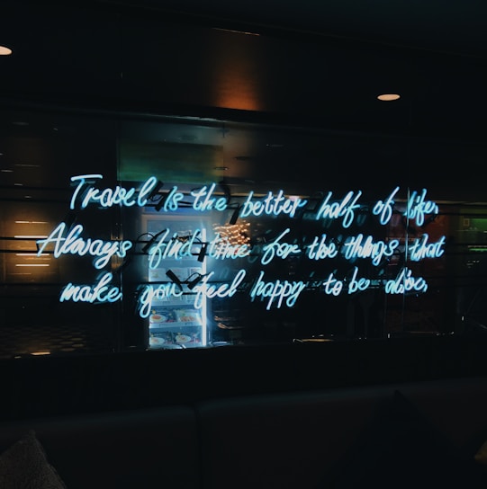 A neon sign with a motivational quote is illuminated against a dark background. The quote encourages taking time for things that bring happiness. Reflections and a slight glow from the neon text are visible.