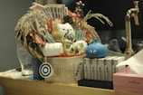A decorative arrangement featuring a variety of items including a golden cat figurine, rice stalks, paper currency, and a blue cartoon-like creature with eyes. The setup is placed on a wooden surface and includes various traditional and cultural symbols, possibly for ornamental or symbolic purposes. A brass faucet is visible in the background, suggesting a kitchen or bar area.