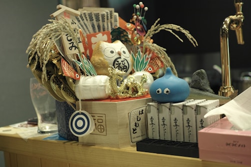 A decorative arrangement featuring a variety of items including a golden cat figurine, rice stalks, paper currency, and a blue cartoon-like creature with eyes. The setup is placed on a wooden surface and includes various traditional and cultural symbols, possibly for ornamental or symbolic purposes. A brass faucet is visible in the background, suggesting a kitchen or bar area.