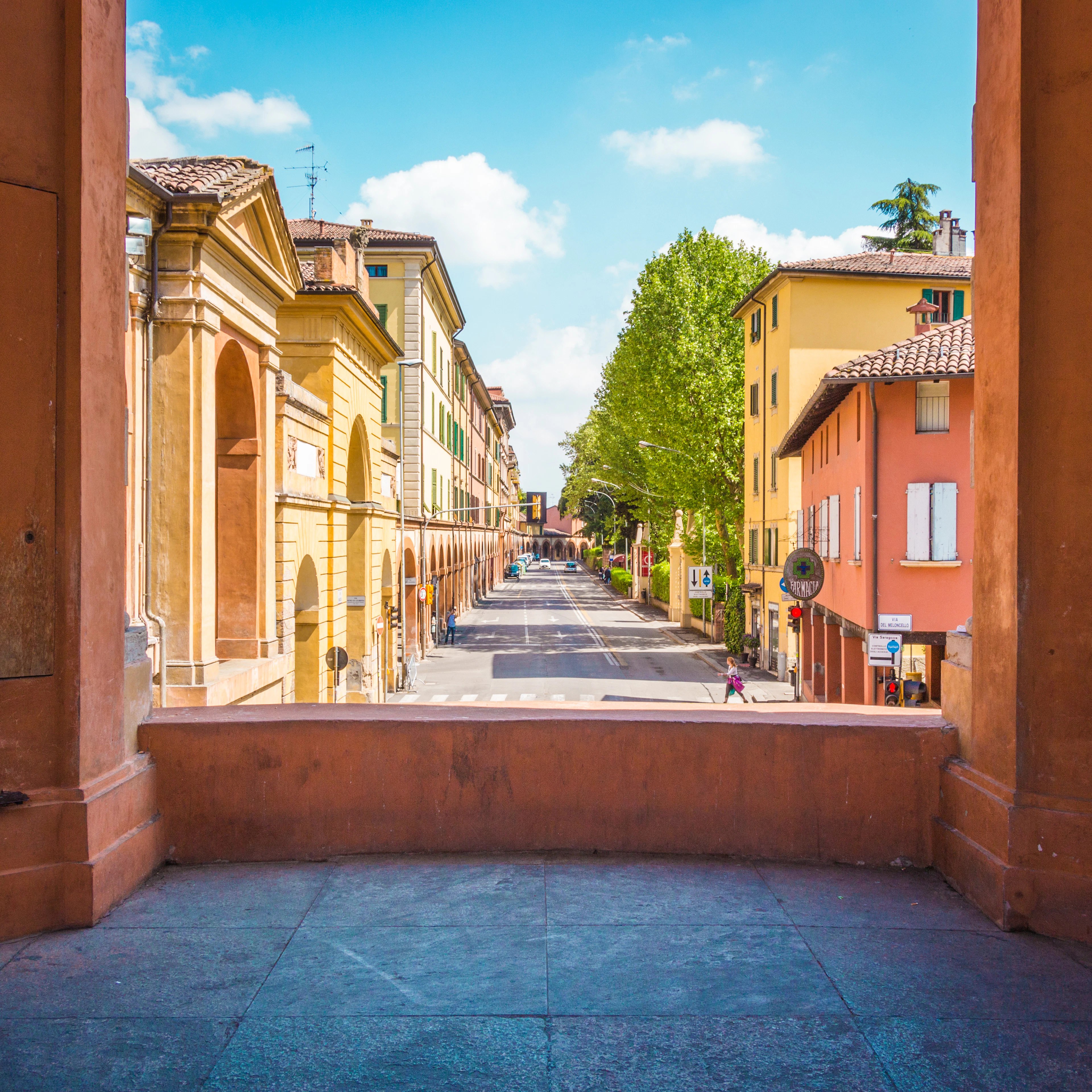 A colonnaded portico frames a sunlit Bologna street lined with ochre and green buildings.