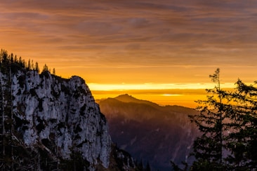 A dramatic sunset over a rugged mountainous landscape. The sky is filled with deep orange and yellow hues, casting a warm glow over the rocky cliffs and dense pine trees in the foreground.