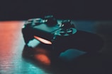 A video game controller is placed on a wooden surface, with soft lighting creating a contrast between the dark device and the background. The lighting casts a warm glow, highlighting the details of the buttons and joystick.