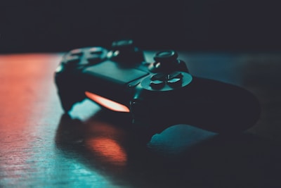 A video game controller is placed on a wooden surface, with soft lighting creating a contrast between the dark device and the background. The lighting casts a warm glow, highlighting the details of the buttons and joystick.