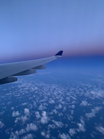 Aerial view showing part of an airplane wing against a stunning backdrop of a clear sky transitioning from deep blue to a purple hue near the horizon. Below, a vast expanse of fluffy white clouds blankets the landscape.