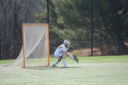 A lacrosse goalie in protective gear stands ready with a stick near the goal on a sports field. The net is clearly outlined in orange, and the goalie appears focused, prepared to block an incoming ball. The background features trees and a fence, indicating an outdoor setting.