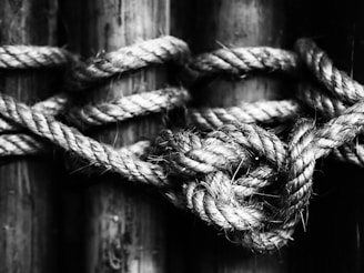 A black and white close-up of a thick rope tied in a knot. The image emphasizes the texture and intricate weaving of the fibers. The background features a wooden surface, creating a rustic and rugged appearance.