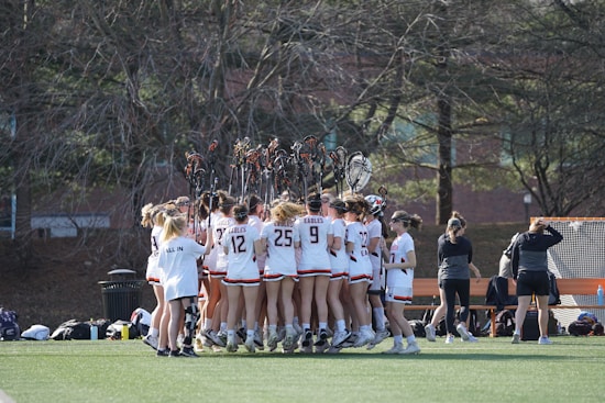 A group of lacrosse players in uniform huddled together on a field, raising their lacrosse sticks in the air. The players are wearing white jerseys with numbers and the word 'Eagles' printed on them. The scene is set outdoors with leafless trees and a building in the background. Various equipment bags and training gear are visible on the ground nearby.