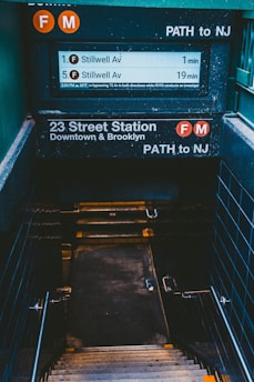 A dimly lit subway station entrance with signage for the F and M trains, indicating directions to Stillwell Avenue and a PATH connection to New Jersey. Stairs descend into the station, surrounded by metal railings and tiled walls.