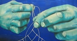 Two large, teal-tinted hands work intricately on assembling or mending a mesh or net with thin, light-colored threads, set against a deep blue background.