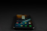 A smartphone displaying various app icons on its screen is placed on a dark surface. The screen is active, showcasing applications like Instagram, Snapchat, and Spotify. The environment is dimly lit, accentuating the glow of the phone screen.
