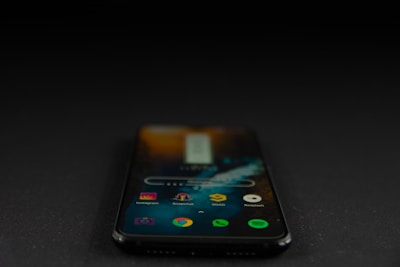 A smartphone displaying various app icons on its screen is placed on a dark surface. The screen is active, showcasing applications like Instagram, Snapchat, and Spotify. The environment is dimly lit, accentuating the glow of the phone screen.