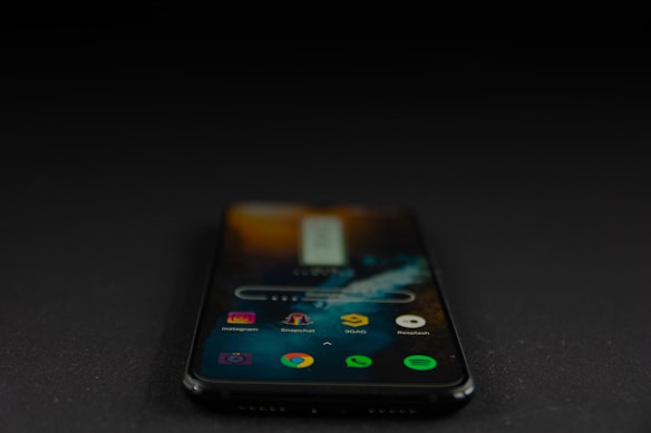 A smartphone displaying various app icons on its screen is placed on a dark surface. The screen is active, showcasing applications like Instagram, Snapchat, and Spotify. The environment is dimly lit, accentuating the glow of the phone screen.