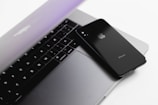 A sleek, modern laptop with a partially closed lid is complemented by a black smartphone placed on top of it. The devices sit on a clean, white surface, showcasing minimalistic and contemporary design.